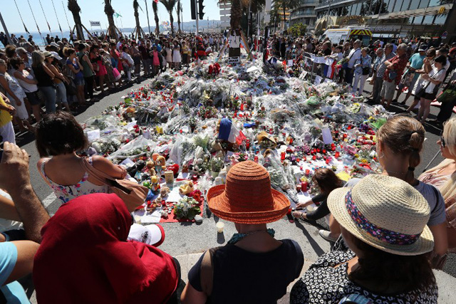 People gather near flowers placed at a makeshift memorial near the Promenade des Anglais in Nice on July 17, 2016, in tribute to the victims of the Bastille Day attack that left 84 dead. The Islamic State group claimed responsibility for the truck attack that killed 84 people in Nice on France's national holiday, a news service affiliated with the jihadists said on July 16. Tunisian Mohamed Lahouaiej-Bouhlel, 31, smashed a 19-tonne truck into a packed crowd of people in the Riviera city celebrating Bastille Day -- France's national day. / AFP PHOTO / Valery HACHE