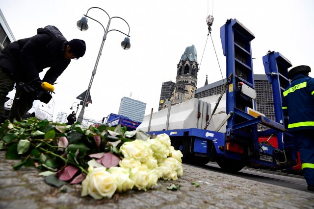 A cameraman films flowers of a makeshift memorial as concrete blocks are put in place with the help of a crane and members of the THW technical relief organisation to secure the Christmas market next to the Kaiser-Wilhelm-Gedaechtniskirche (Kaiser Wilhelm Memorial Church) on December 22, 2016 in Berlin, three days after a truck crashed into the market, killing 12 people and wounding almost 50.. The Berlin Christmas market that was struck by a deadly truck rampage reopens on  December 22, 2016, organisers said, as the grieving city sought a return to normal life and police hunted for the prime suspect in the attack. / AFP PHOTO / Tobias SCHWARZ